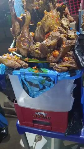 Here is a description of the video:

The video shows a street food vendor in what appears to be an outdoor setting. The focus is on a large pile of cooked and seasoned meat, likely chicken or poultry parts, stacked on a platter. The platter is placed on top of a white plastic container, which itself sits on a red crate labeled "AMSTEL Malta". The meat is visibly garnished with chopped red and green peppers. A hand wearing colorful beaded bracelets briefly reaches into frame, possibly belonging to the vendor. The background includes other objects, possibly part of the vendor's stall setup and other boxes or crates nearby. The lighting suggests it's evening or night, and there seems to be some activity around the stall.