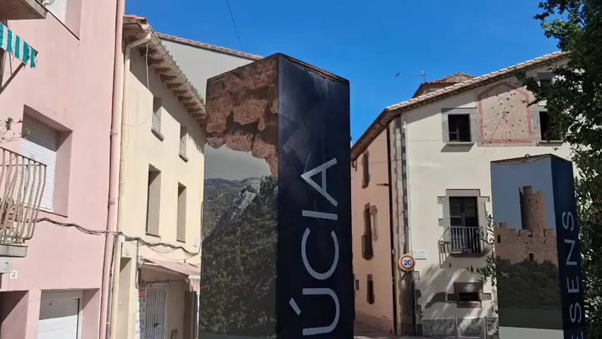 The video shows a street in the town of Sant Julià de Cerdanyola, Spain. The street is lined with two-story buildings painted in light colors. On either side of the street are tall, rectangular signs with pictures and text about the town. The sign on the left has the name "SANT JULIÀ," written in white, vertically on a black background. The sign on the right says "ROQUESENS" in the same style.  A speed limit sign is visible on the right, and trees are visible in the background.