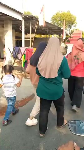 The video shows a group of people walking down a paved street. In the foreground, a young child with pigtails, wearing a striped shirt and jeans, is walking and holding hands with an unseen person. Behind the child, two women in hijabs walk, one in a brown hoodie and the other in a light brown hijab and a teal shirt. Another person in a red top is partially visible. Further back, a man in a blue shirt and black pants, and another man in a black t-shirt with an ornate design on the back and a black baseball cap are walking. A clothesline with various garments is visible on the left side of the street, along with some buildings and trees. Two Indonesian flags are prominently displayed on poles. The scene appears to be outdoors in daylight. The camera is mounted on a vehicle, likely a motorcycle, as indicated by the handlebar visible at the bottom of the frame.