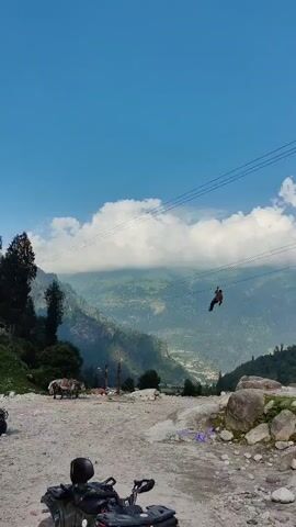 A person is zip-lining across a mountainous landscape. The person is suspended on a wire, with a steep drop below. The background is filled with lush green mountains and a cloudy sky. In the foreground, there is a gravelly surface with some rocks and vegetation. An ATV is parked on the left side of the frame, and a donkey is standing nearby.