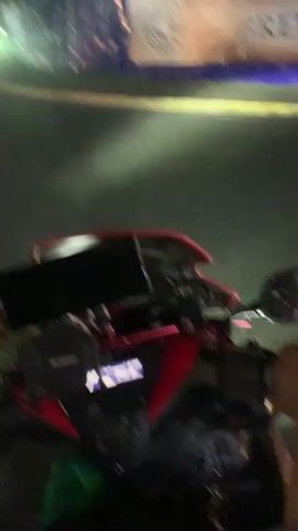 This is a first-person perspective video of someone riding a red motorcycle at night in Angeles, Philippines. The rider's hands are visible on the handlebars, and the motorcycle's digital dashboard is in focus, displaying the time as 6:09 and a speed of 29 km/h. The scene is a street at night, illuminated by streetlights and the headlights of passing vehicles. 

In the background, there are buildings, including a modern white structure on the left. Further down the road, a large truck and other vehicles are visible. A blue construction barrier with signage can be seen on the right side of the road. The road appears to have a pedestrian crossing with thick white stripes. The overall mood is that of a nighttime ride through a town. There is no discernible weather information, but the darkness and artificial lighting suggest it is nighttime. The visible text on the dashboard includes "MOTOWOLF" and "6:09" and "29 km/h". There is also text on the signage in the background, but it is too blurry to read.