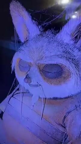 The video shows a life-size, somewhat dilapidated model of Po, the panda from Kung Fu Panda, sitting in a position, possibly on the ground, or on a seat. His eyes are closed. The background suggests a possibly theatrical, constructed environment, illuminated with blue light.  A man in glasses briefly appears in the frame.