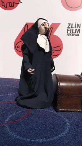 A full shot depicts a figurine of a nun standing next to a small, dark wooden chest. Behind the nun and chest is a white backdrop displaying the logo for the Zlín Film Festival. The nun figurine is dressed in a long, black habit with a white wimple and a silver cross pendant. The wooden chest has a curved lid and is detailed with metallic studs. The figurine and chest are positioned on a blue carpet with a circular pattern of red lines.  The scene appears to be set up for display or promotional purposes, possibly at the film festival itself.