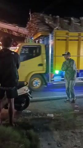 The video shows a nighttime scene on a street, possibly in a rural or suburban area. A large yellow truck carrying a load of what appears to be wood or building materials is driving by. The truck has colorful decorations and lights. In the foreground, there are two young men standing near a parked scooter. One man, wearing a black jacket and shorts, is partially visible on the left. The other man, wearing a grey hoodie and jeans, is facing the street, observing the truck. Several other people and vehicles, including motorcycles and a white car, are visible in the background. There are also what appear to be pieces of trash on the ground. The overall atmosphere seems to be casual, with people gathered to watch the passing truck. There are Indonesian flags visible on some of the buildings.