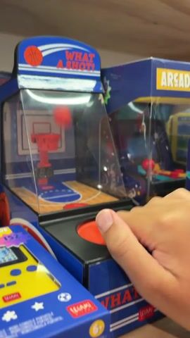 A close-up shot shows a person's hand playing a miniature basketball arcade game. The game, labeled "WHAT A SHOT!", features a small basketball court, a hoop, and a red basketball. The person is pressing a large red button on the front of the game. The basketball bounces within the game, and a digital display shows the score. The game is surrounded by other similar arcade game boxes. The overall environment appears to be a store display.