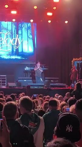A man is singing on a stage during a concert. He is wearing a gray shirt and jeans, and holding a microphone. Behind him are two band members and a large screen that says "Melody." The stage is lit up with red and white lights. In front of the stage is a crowd of people, many of whom are holding up their phones to record the performance. Some of the people in the crowd are wearing green scarves.