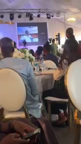 The video shows an event in a room with tables and chairs set up. There are people seated around the tables, some looking at a large screen that is displaying a video. The video appears to be a presentation, possibly celebrating an anniversary or a special occasion. A person is standing near the tables, interacting with others. The room is well-lit with stage lights and decorated with white flowers. The overall atmosphere seems formal and celebratory.