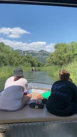 Two people are seated in the back of a boat, facing away from the camera and looking forward. The person on the left is wearing a light pink t-shirt and a dark bucket hat. The person on the right is wearing a black t-shirt and has short, light brown hair. They are on a boat traveling down a narrow river or canal. The water is calm and green, reflecting the blue sky and white clouds. Lush green vegetation, including reeds and trees, lines both sides of the waterway. In the distance, a larger boat with at least two people on board is also moving forward. Beyond the trees, a large, rocky mountain range is visible under a bright, sunny sky. The overall scene suggests a leisurely boat ride on a warm, clear day, possibly a tour or a recreational outing. The visible text on the t-shirt of the person on the right appears to be a stylized logo, possibly an animal silhouette. The time of day appears to be midday, given the bright sunlight and the shadows. The location is Prevlaka, Montenegro, suggesting a scenic natural environment, perhaps a national park or a protected area known for its waterways and mountainous scenery.