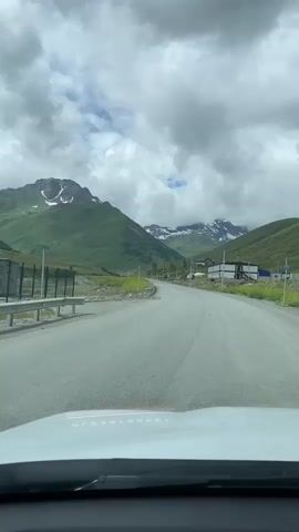 The video shows a point of view shot from inside a car driving on a paved road. The road is surrounded by lush green mountains and a cloudy sky. The mountains have patches of snow on their peaks. There is a guardrail along the side of the road, and some buildings are visible in the distance. The car appears to be moving at a moderate speed.