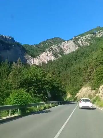 The video shows a white car driving on a paved road through a mountainous, forested area on a clear day. The road curves to the right. The car is in the right lane. To the left of the road is a metal guardrail and dense green trees. To the right of the road, there is a steep, rocky embankment covered in trees and vegetation. In the background, there are large, sheer rock faces and dense green forests covering the mountains. The sky is a clear, bright blue. The camera appears to be mounted inside another vehicle, possibly following the white car.