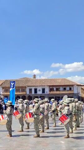In a vertical shot, a line of drummers stands on a brick surface, playing their instruments. They are positioned in front of a building, with a crowd of people standing on steps leading to the building. Banners are displayed in front of the building. The sky is blue with some clouds. The drummers are dressed in military style uniforms. They are playing red drums. The crowd is a mix of people wearing a variety of clothing.
