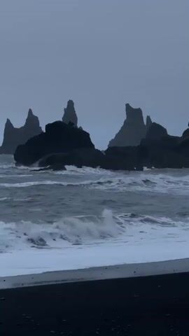 The video shows a seascape with dark, jagged rock formations emerging from the ocean under a cloudy sky. Waves break against the rocks and wash onto a black sand beach in the foreground. The overall mood is somber and dramatic. There are no people visible.