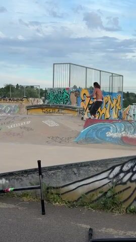 The video shows a man riding a scooter in a skatepark. The man is shirtless, wearing dark pants and sneakers. He has long hair. He rides over a ledge, then rides along a sloped side. Another boy is riding a scooter on the other side. The skatepark has various slopes and ledges, covered in graffiti. A fallen scooter lays on the ground. A chain-link fence is visible in the background. The sky is overcast.