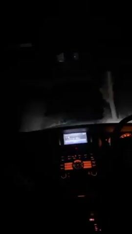 The interior of a car is shown at night, with the headlights illuminating a dark, winding road ahead. Trees line the sides of the road. The car's dashboard is visible, showing the radio display. A child's voice is audible.