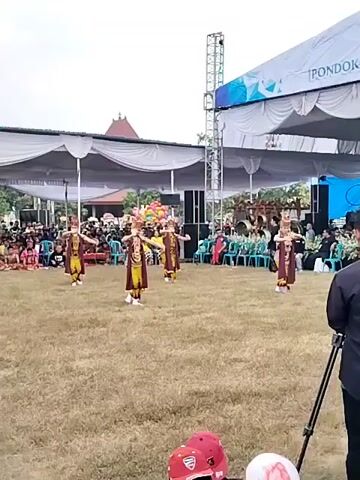 A group of five young people, dressed in traditional ethnic clothing with elaborate headwear, are performing a dance on a dry, grassy field. They are spread out in a scattered formation across the field. In the background, there are tents and a stage setup with a large screen displaying graphics. An audience is seated behind the performers, under the tents, and more people are standing. A tripod with a camera is visible in the foreground. The event appears to be an outdoor cultural performance.