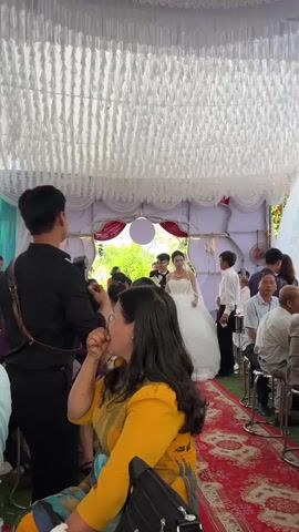 The video captures a wedding reception or ceremony entrance taking place under a large, decorated tent in Bình An, Vietnam.

The main subjects are the bride and groom, who are walking down a central aisle. The bride is dressed in a traditional white, floor-length wedding gown with intricate embellishments on the bodice, a full skirt, and a veil. She wears a gold necklace and bracelet. The groom, walking beside her, is in a black suit with a white shirt and black bow tie. Both appear to be smiling.

Numerous guests are seated at round tables on either side of the red carpeted aisle, watching the couple. The tables are set with drinks and possibly food, and guests are seated on stools. In the foreground, a woman with long dark hair, wearing a bright yellow top and patterned trousers, is seated with her back mostly to the camera, looking towards the approaching couple. A black handbag rests on her lap.

The tent's ceiling is adorned with white, ruffled fabric drapes, creating an elegant atmosphere. The background features a decorative archway, possibly marking the entrance to the reception area, with a large white balloon or spherical decoration visible through it. Beyond the tent, glimpses of natural light and green foliage suggest an outdoor or semi-outdoor setting during daylight hours. The overall scene conveys a festive and celebratory occasion. No explicit text is visible in the frame.