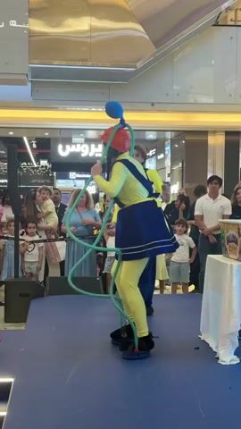 A performer dressed in a yellow jumpsuit and blue skirt, wearing a red hat and black shoes, is balancing on a blue platform. They are holding a green tube that loops around their body, with a blue balloon attached to the top. The performer is standing on a blue stage in front of an audience of children and adults. In the background, there are shops and escalators inside a mall. The performer is looking up and to the right, possibly at the balloon. Another performer in a yellow top is partially visible behind the main performer. The scene is well-lit with artificial lighting.