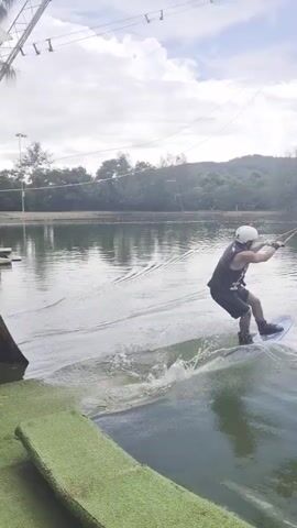 This media file captures a wakeboarder in action at a cable park in Tambon Mai Khao, Thailand. The scene is set on a calm body of water, likely a lake or pond designed for wakeboarding, surrounded by lush green trees and a cloudy sky. The time of day appears to be daytime, with the overcast weather casting a diffused light on the scene.

The primary subject is a wakeboarder, seen wearing a helmet and dark clothing, actively engaged in riding the wake. The wakeboarder is being pulled by a cable system, evident from the overhead wires and pulley mechanisms visible in the background. This cable system suggests a controlled environment for wakeboarding, common in dedicated parks.

The wakeboarder is creating a significant splash as they carve through the water, demonstrating skill and speed. The water itself is choppy in the wakeboarder's path, contrasting with the smoother surface elsewhere. In the distance, there are structures that appear to be part of the wakeboarding facility, possibly ramps or obstacles. The overall impression is one of an active recreational pursuit taking place in a scenic, natural setting. There is no visible text in the media file.