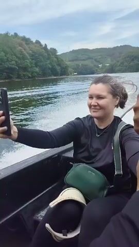 A boat ride on a lake with lush green hills on either side. The boat appears to be moving at a moderate speed, creating a wake behind it. The sky is partly cloudy. In the foreground, a woman is taking a selfie with her phone. She is wearing a black long-sleeved shirt and has a green crossbody bag. To her right, a person's hands are visible gripping a metal pole. In the background, a child wearing a blue hat and a rash guard is seated and holding a dark-colored bottle with a straw. Further in the distance, there are buildings and other boats on the water. The video also shows a brief glimpse of the boat's canopy and the surrounding scenery.