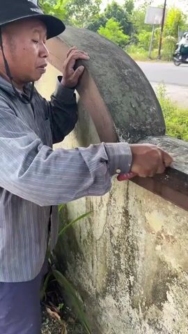A person wearing a black hat and a striped shirt is shown from the side. They are holding a small object, possibly a knife, and appear to be scraping or cleaning a wooden surface that is attached to a concrete structure. The concrete structure is weathered and appears to be part of a wall or a building. The background shows a road, greenery, and a white fence. The lighting suggests it is daytime.