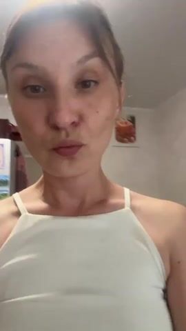The video shows a woman with her hair pulled back, wearing a white tank top. She is facing the camera and appears to be in a room with artwork on the walls and a microwave visible in the background. She makes a few facial expressions and moves her head.