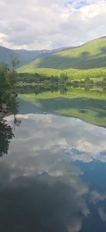 The video shows a reflection of a lush green mountain range on a calm body of water. The sky above is partly cloudy, with some sunlight casting shadows on the mountains. The water's surface is smooth, creating a clear mirror image of the landscape. The scene is tranquil and scenic, typical of a natural outdoor environment.