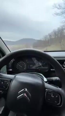 The video shows a view from inside a car, focusing on the steering wheel, dashboard, and the road ahead. The car, a Citroen as indicated by the logo on the steering wheel, is traveling on a slightly winding road under a cloudy sky. The landscape outside consists of trees and a distant hill. The speedometer reads approximately 55 km/h. The driver's left hand is visible on the steering wheel. A smartphone is mounted to the left of the steering wheel.  Music is playing in the background.