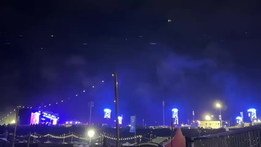 The video shows a wide outdoor night shot of a festival or concert grounds. In the foreground, there are string lights hanging between wooden poles, creating a festive atmosphere. Further back, a large stage with bright lights is visible, suggesting a performance is underway. To the right of the stage, multiple tall screens display blue lights. The sky is dark, but some green lights are visible, possibly from drones or lasers. Wisps of smoke or fog drift across the scene, illuminated by the various lights. The overall impression is a lively outdoor event at night.