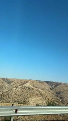 The video shows a landscape view from a vehicle, likely looking out a window. The foreground features a silver metal guardrail and some dry vegetation. Beyond the guardrail, a vast expanse of arid, rolling hills dominates the scene. The hills are covered in dry brush and sparse vegetation, with some areas appearing eroded or terraced. In the distance, more hills and a hazy horizon are visible. The sky is a clear, bright blue with a faint contrail from an airplane visible in the upper right portion of the frame. There are no people or discernible objects in motion within the landscape itself.