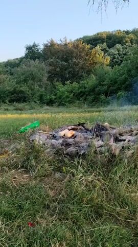 The video pans across a grassy, outdoor scene. In the foreground, a camouflage backpack is visible, with a flashlight attached to it. Several white paper items are scattered on the grass near the backpack. Further into the scene, a picnic blanket is laid out with a bottle of water and some food items. Beyond that, a campfire is situated in a clearing, with some logs and ashes visible. In the background, a lush, green hillside covered in trees and foliage rises under a clear sky. The lighting suggests it is either early morning or late afternoon.