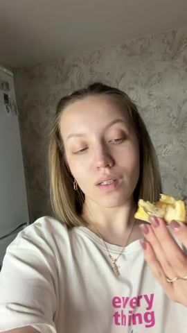 A young woman is taking a selfie video while eating. She holds a partially eaten piece of food with her left hand, bringing it up to her mouth to take a bite. She chews with her mouth closed and swallows, looking up slightly before looking directly at the camera. The woman has light brown hair, light skin, and blue eyes. She is wearing a white t-shirt with pink text. She has a gold cross necklace and gold earrings. She is also wearing a gold ring. The background is a patterned wall and a white refrigerator is partially visible on the left. The camera angle is slightly downward.