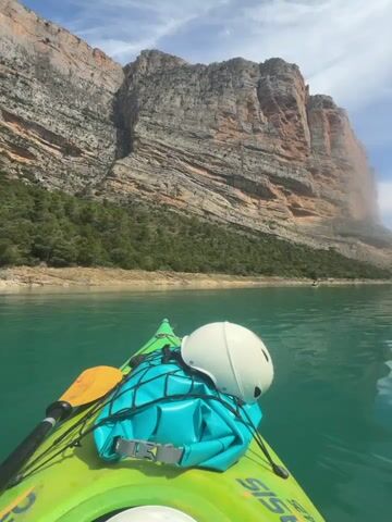 A first-person view from a bright green kayak shows a person kayaking on a calm body of turquoise water. The kayak has a white helmet and a teal dry bag strapped to its front. Steep, rocky cliffs rise on both sides of the water, with some greenery visible at their bases. The sky is partly cloudy with patches of blue. In the distance, other kayakers can be seen. The camera slowly pans from left to right, revealing more of the scenic canyon and water.