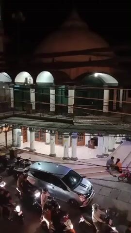 The video captures a night view of a mosque with a large dome and a tall minaret. The mosque is illuminated, highlighting its architectural features. In the foreground, a silver minivan is parked, with several motorcycles also visible. A few people are seen near the entrance of the mosque, some sitting on the steps and others standing. The scene suggests a public area, possibly a town square or marketplace, adjacent to the mosque. The lighting is artificial, indicating it is nighttime. The overall atmosphere appears calm and serene.