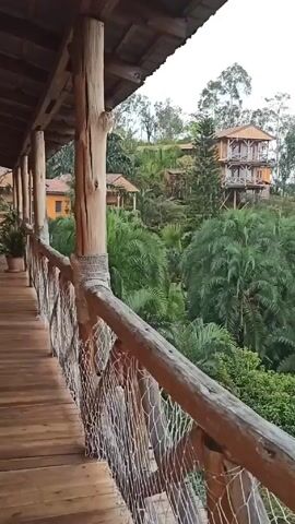 The video shows an elevated walkway with a wooden railing, leading to a view of several buildings on a hillside covered in lush green vegetation. The walkway itself is made of wooden planks, supported by wooden posts wrapped with rope-like material. The railing consists of wooden beams and a woven net-like structure. In the distance, several buildings are visible, including a multi-story structure built on stilts and other single-story buildings with orange walls and red tile roofs. The hillside is densely populated with trees, including palm trees and taller evergreens. The sky is overcast, and it appears to be raining lightly.