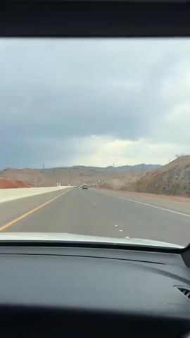 The video captures a perspective from inside a vehicle driving on a highway. The scene is a desert landscape with rolling hills and mountains in the background, under a cloudy, overcast sky. The road is asphalt with clear lane markings and a yellow line on the left. Several other vehicles, including cars and trucks, are visible on the highway ahead and in the distance. Power lines and poles are present throughout the landscape, indicating infrastructure. The overall atmosphere appears somewhat muted due to the cloudy weather, suggesting it might be a cooler part of the day or a storm is approaching. There is no visible text in the video. The location context provided places the scene in Boulder City, USA.