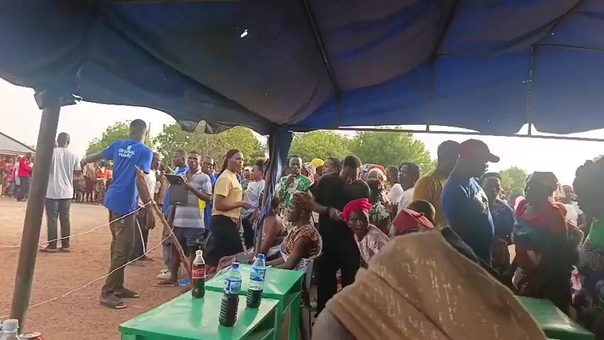 The video shows a gathering of people in an outdoor area. There is a tent set up with tables covered in green, and bottles of water and soda are visible on the tables. People are standing under the tent and around it. Outside the tent, there is a roped-off area where a person dressed in a traditional beige robe is walking. A large crowd is watching from behind the rope, and there are several buildings in the background, possibly school buildings. Several people are wearing blue t-shirts with the words "G-K-O-G" printed on them, and some people are sitting on chairs behind the tables. The event appears to be some kind of outdoor celebration or community gathering.
