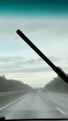 The video depicts a driver's perspective while traveling on a wet, possibly rainy, highway. The windshield is covered with raindrops, and windshield wipers intermittently clear the view. The road is a multi-lane highway with visible lane markings. Other vehicles, including cars and what appear to be trucks, can be seen in the distance ahead. Trees line the sides of the road, suggesting a rural or semi-rural environment. The sky is overcast with gray clouds. The general atmosphere is that of a potentially gloomy or dull day due to the weather conditions.