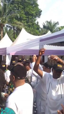 The video captures a gathering of people, mostly men, outdoors under white tents with light purple trim. Many attendees are wearing white shirts and black baseball caps with gold writing, while others sport colorful African-patterned clothing. Some people are raising their arms, with money visible in their hands. The background features palm trees and a building. It appears to be a celebratory event or a political rally.