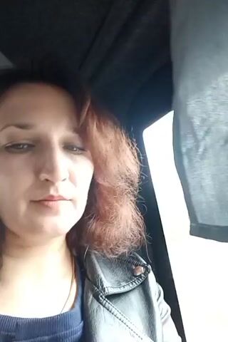 A woman with auburn hair and wearing a black leather jacket sits in a car. She is looking at the camera and smiling slightly. Outside the car window, green trees and a road can be seen. The car is moving. A grey fabric curtain is partially drawn over the window frame.