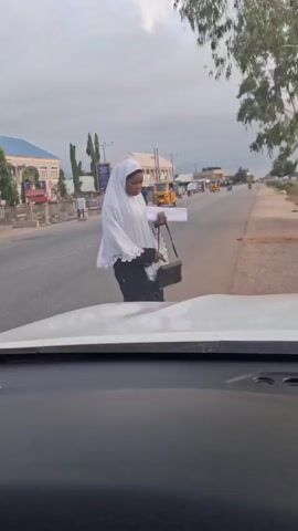 From inside a moving vehicle, a person in a white head covering, dark top, and carrying papers is walking on the side of a road. In the background there are buildings, a tuk-tuk, and people. As the car moves, the surroundings change, and the woman remains walking on the side of the road. The background transitions to a construction site and a dirt road. A person can be seen walking in the distance.