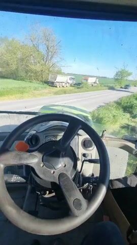 The video shows a view from inside a tractor cabin, looking out onto a rural scene. The tractor's steering wheel and dashboard are visible in the foreground. Through the windshield, there is a straight paved road with green grass on both sides. On the other side of the road, two large white dump trucks are parked. A black car passes by on the road. The sky is clear and blue. The video then pans upwards, showing the interior of the cabin more clearly, including a rearview mirror which reflects the face of the person recording the video.