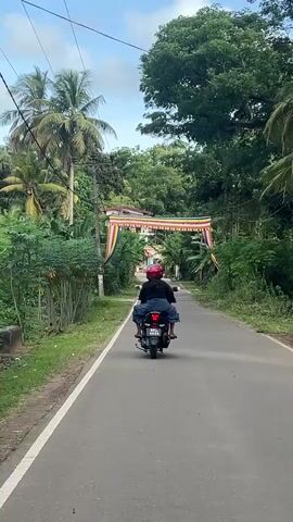 A motorcycle carrying two people travels on a narrow asphalt road in a rural, tropical setting. The motorcycle is seen from the rear, with the rider wearing a black shirt and the passenger behind wearing blue clothing and a red helmet. Lush, green vegetation flanks both sides of the road, including tall trees and various plants, creating a dense canopy in places. Overhead utility lines run parallel to the road. The motorcycle passes under a large decorative arch or gateway adorned with colorful Buddhist flags and banners. The sky above is a light blue with scattered white clouds.