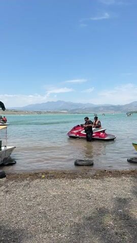 Two people are on a red jet ski in the water near the shore. The person in the back is wearing an orange life vest. The person in the front is wearing sunglasses and a dark shirt. The water is a light blue-green color. In the background, there are mountains and a clear blue sky with some clouds. There are also other boats and people in the water further out. The shore in the foreground is covered in pebbles and gravel, with some tires and a yellow and green boat on the right.