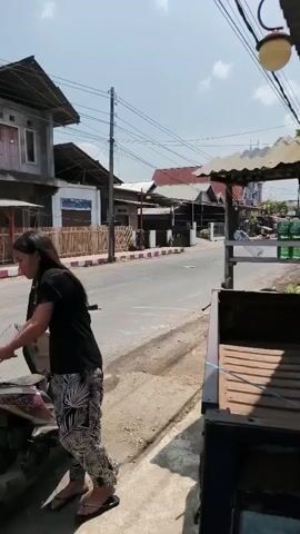 The video shows two people interacting near a parked motorcycle on a street. A man wearing a black long-sleeved shirt and dark pants stands to the left of the motorcycle, facing it and holding a yellow bowl. A woman wearing a black t-shirt and patterned pants stands to the right of the motorcycle, also facing it. She is holding a clear plastic bottle and appears to be pouring liquid from it into the yellow bowl held by the man. The motorcycle is a scooter-type, with its seat open. The background features buildings with visible windows and roofs, a street, and a clear sky. Power lines are visible above the buildings. The lighting suggests it is daytime, likely sunny.