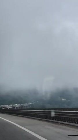 The video captures a view from a moving vehicle on a highway in Churek, Bulgaria. The scene is dominated by a thick layer of fog or low clouds that obscure the upper portions of the surrounding mountains and a large bridge. The bridge appears to be a viaduct, supported by numerous pillars, and carries traffic. Several vehicles, including what seem to be trucks and cars, are visible on the bridge, moving slowly or stopped, suggesting potential traffic congestion.

The weather is overcast and misty, contributing to the subdued atmosphere and limited visibility. The highway itself is paved, with clear lane markings, including a solid white line. A metal guardrail runs along the side of the road. On the left side of the road, there are trees and vegetation, and a tall electricity pylon is visible, standing out against the foggy backdrop. A billboard is also partially visible on the left.

The overall mood is one of travel through a somewhat somber, possibly damp, mountain landscape. The limited visibility and the presence of traffic on the bridge create a sense of a journey being undertaken despite the challenging weather conditions. No people are distinctly visible as subjects within the frame. The time of day is not explicitly clear, but the overcast conditions suggest it is daytime.