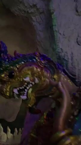 A dimly lit, cave-like environment features a large, ornate statue of a mythical creature. The statue appears to be made of a metallic material, possibly bronze or gold, and is richly decorated with colorful accents of green, purple, and blue. The creature has a fierce, dragon-like head with sharp teeth and elaborate headdress. Its body is depicted as serpentine, with scales that shimmer under the lighting. The statue is partially integrated with the rough, stone-like walls of the cave, suggesting it's part of the natural structure or an elaborate temple setting. The lighting is dramatic, with colored spotlights highlighting the statue and casting shadows, creating an atmospheric and potentially mystical ambiance. The camera pans slowly, capturing different angles of the statue and its surroundings.
