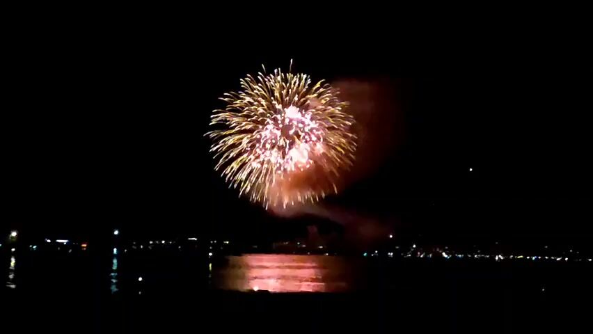 The video shows a fireworks display over a body of water at night. The fireworks are a mix of yellow, pink, and white, and they are exploding in a spherical pattern. The reflections of the fireworks are visible on the water. The shoreline in the background is silhouetted against the bright lights of what appears to be a city. The sky is dark, with a few faint stars visible. The overall impression is one of a celebratory and festive atmosphere.