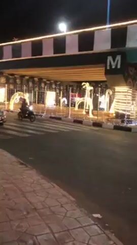 The video shows a street at night that is decorated with holiday lights. A black motorcycle with a rider goes by, followed by a silver sedan. The road goes under a bridge decorated with white and yellow lights. The sidewalk is brick, and a streetlamp is visible.