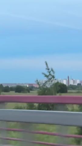 The video shows a blurred landscape seen from a moving vehicle. In the foreground, there are blurred railings of a bridge or overpass, with a red and white stripe visible on the top railing. Beyond the railing is a field with grass and scattered trees, some of which are blurred by motion. In the distance, a long, light-colored multi-story building with many windows is visible, along with a tall, reddish-brown industrial chimney. The sky is a pale blue with some lighter streaks. The overall impression is that of a daytime scene viewed from a moving vehicle on a highway or elevated road.