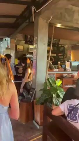 The video shows a woman in a blue dress walking a black and white dachshund on a tiled surface in what appears to be a restaurant or bar. The lighting is bright and sunny, and there are other people and objects like potted plants visible in the background. The dog is walking ahead of the woman.